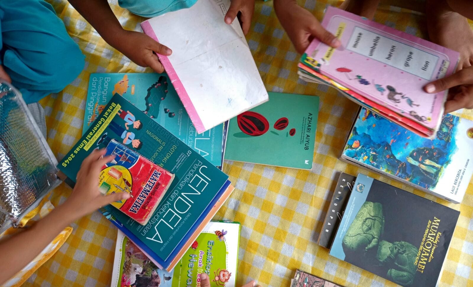 Tangan anak-anak sedang membuka dan membaca buku pelajaran di atas tikar
