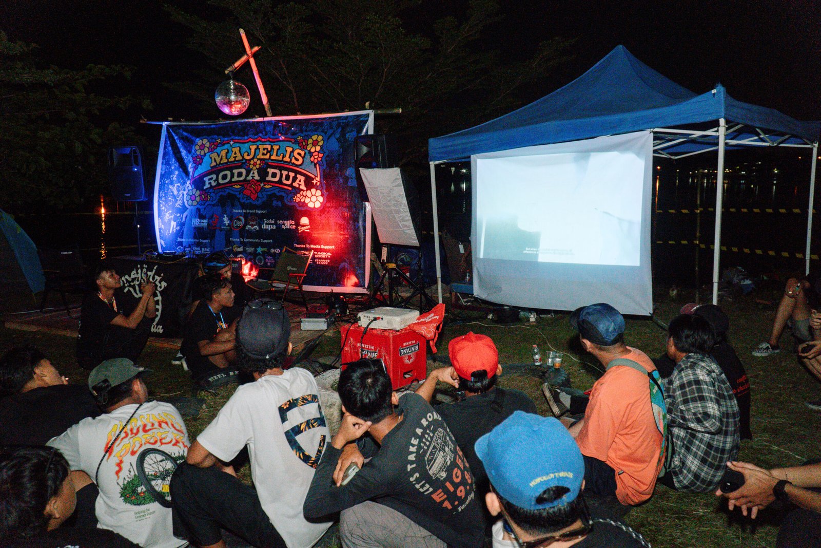 Peserta duduk bersama menyaksikan pemutaran film outdoor pada acara Majelis Roda Dua di tepi danau pada malam hari.